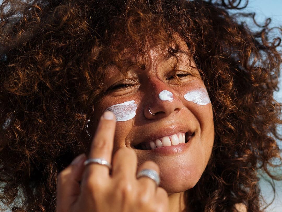 Young woman with curly hair and light brown skin applying sunscreen on her cheeks and nose Desktop
