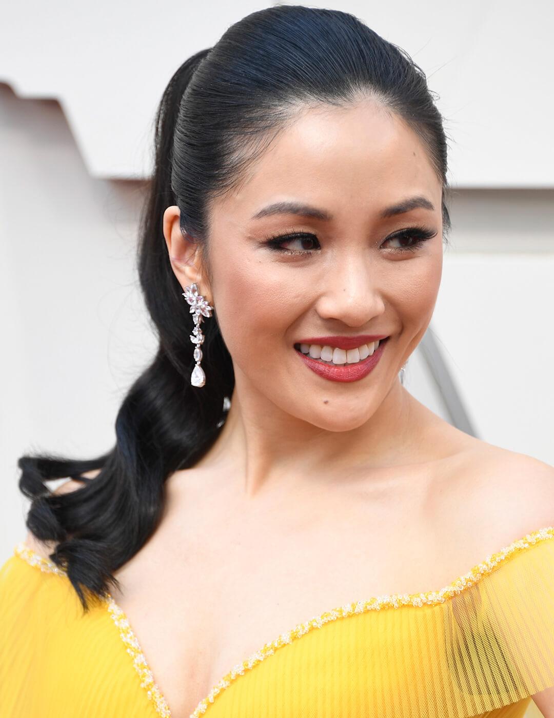 Constance Wu looking regal in a yellow dress, wavy ponytail hairstyle, and silver dangling earrings Desktop