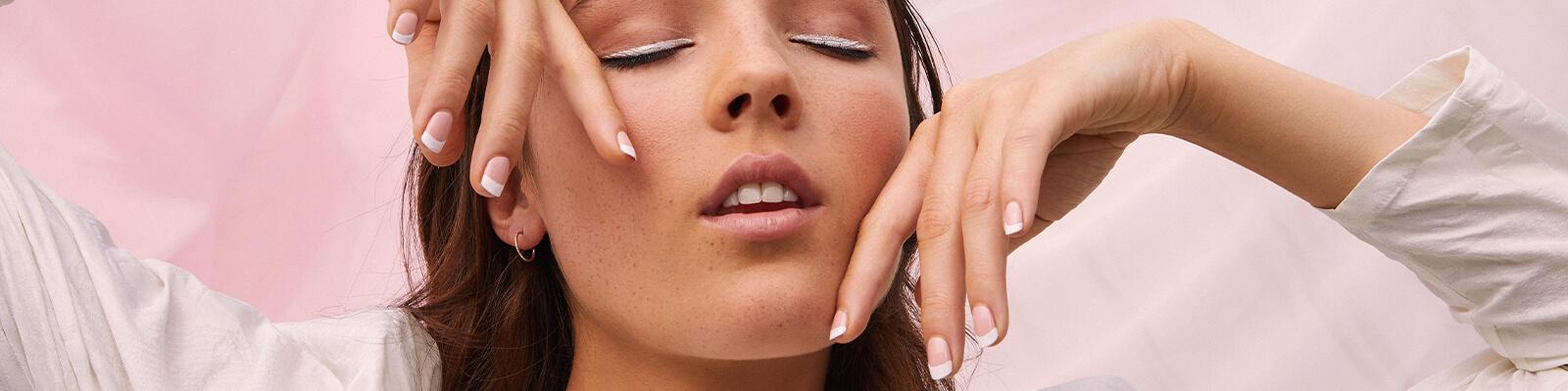 A closeup image of a model with a white eyeliner look and French tip mani posing with her two hands