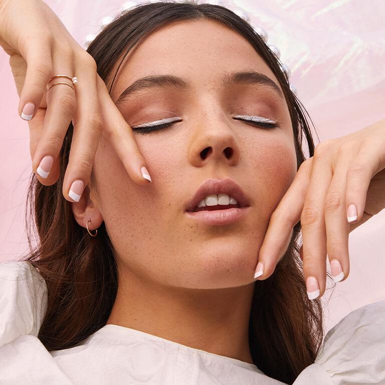 A closeup image of a model with a white eyeliner look and French tip mani posing with her two hands