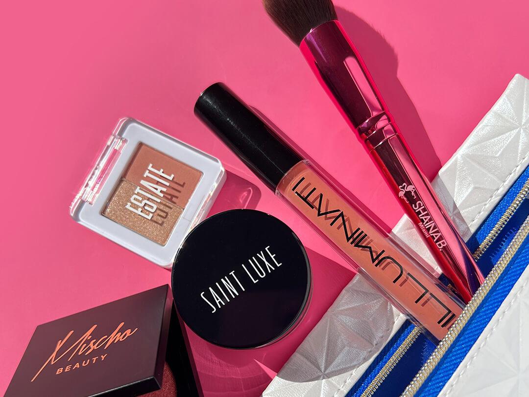 Makeup products and tools from various brands and small silver pouch on dark pink background Desktop