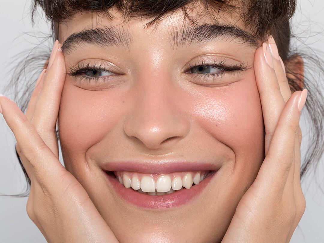 Close-up image of a woman with clear, smooth skin smiling while touching her face Desktop