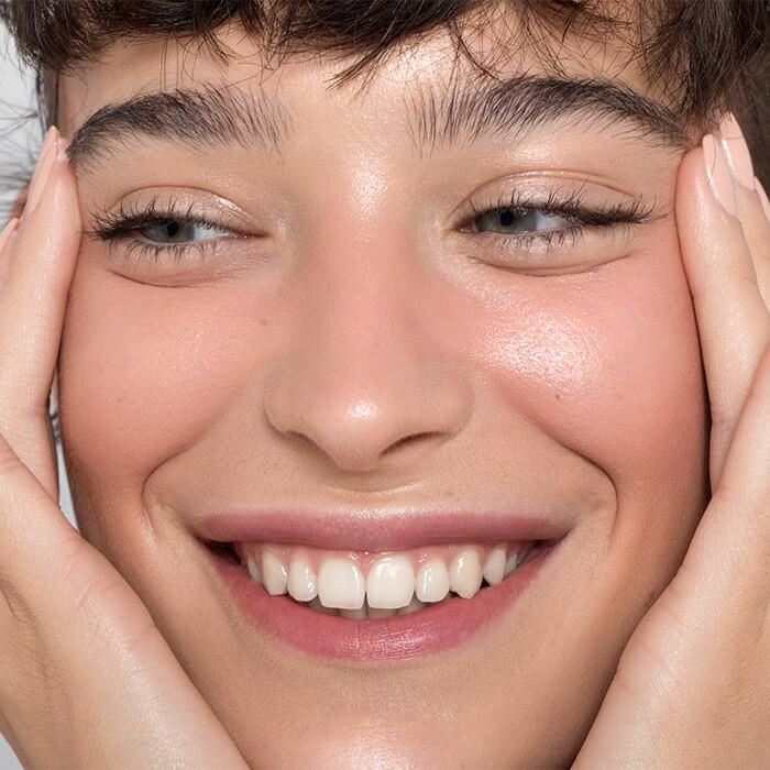 Close-up image of a woman with clear, smooth skin smiling while touching her face Mobile