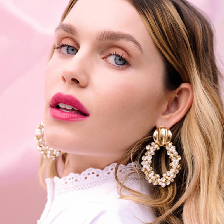 A closeup image of a model wearing nude eyeshadow and bold pink lipstick and gold and pearl hoop earrings Desktop
