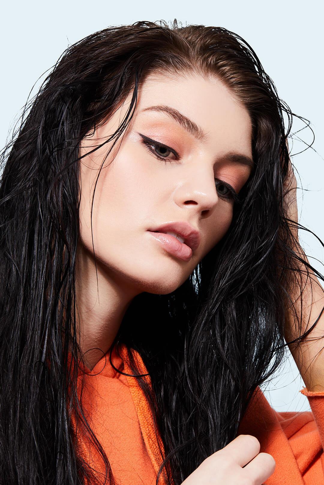 An image of a woman who has long, black hair with her gaze directed downwards dressed in an orange top and wearing a light makeup with eyeliner Desktop