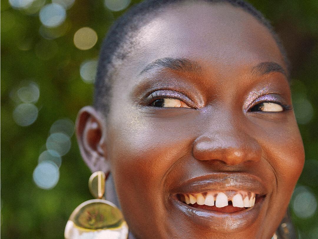 Image of a model with a shimmery purple eyeshadow look and bold statement earrings smiling big while looking to the side Desktop