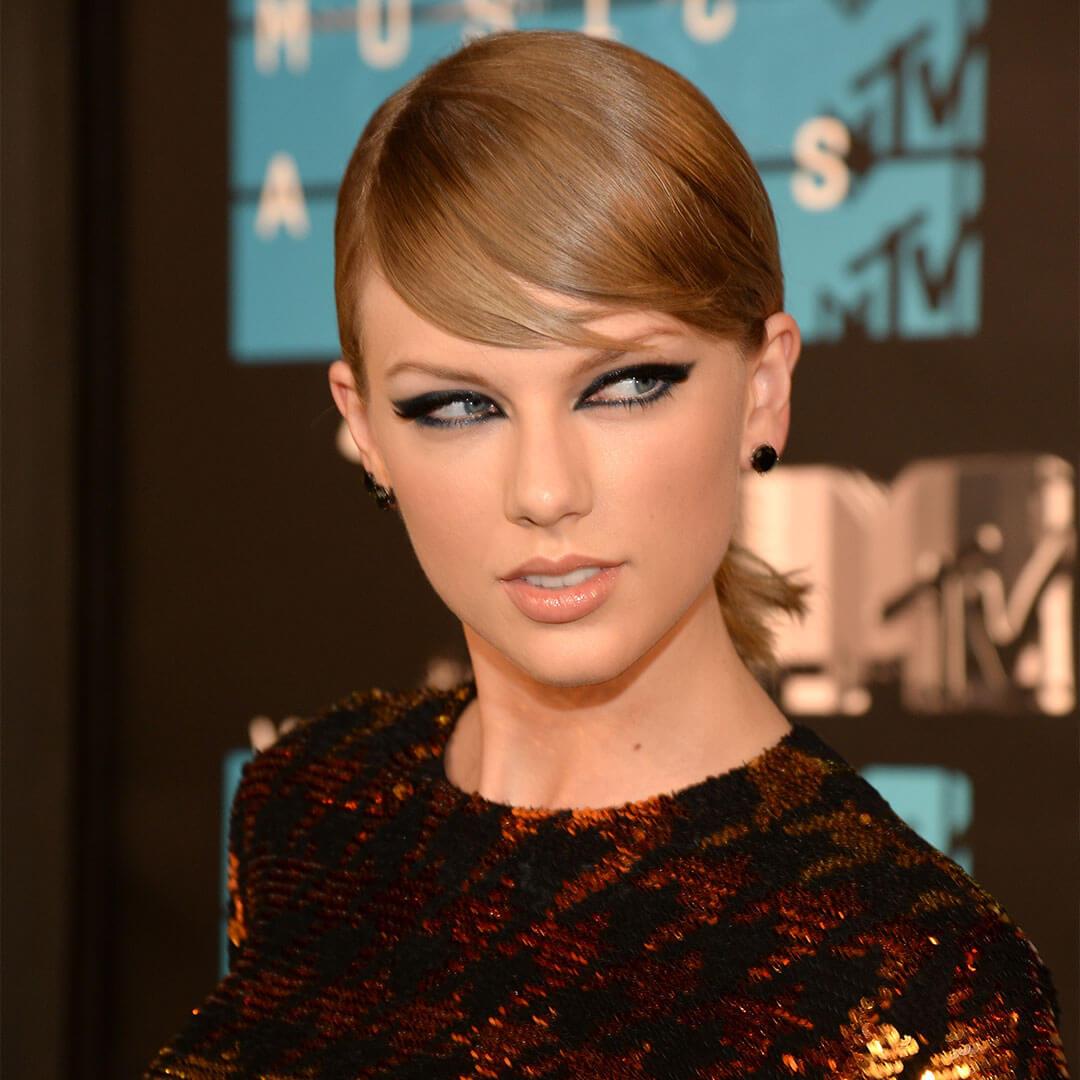An image of Taylor Swift featuring bold eyeliner and nude lipstick, with her hair styled in slick, sideways bangs, wearing black earrings and a fitted long-sleeve top adorned with sequins Desktop