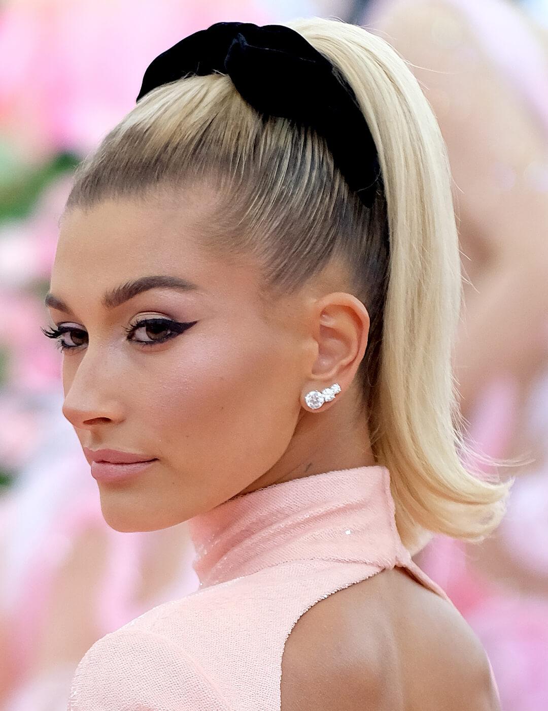 Hailey Bieber looking chic in a baby pink dress paired with a bold eyeliner look and a high ponytail hairstyle kept together with a black velvet scrunchie Mobile