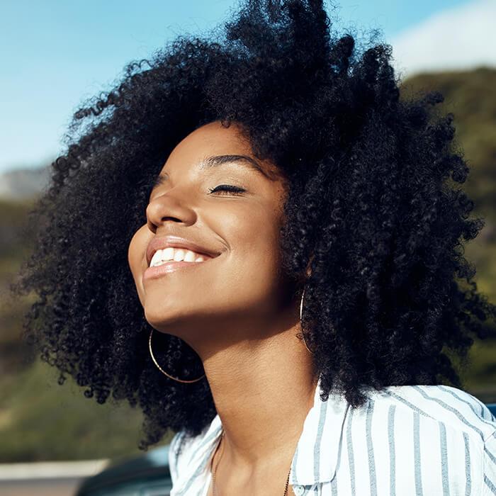 Close-up of a woman of color smiling in the sun against greenery background Mobile