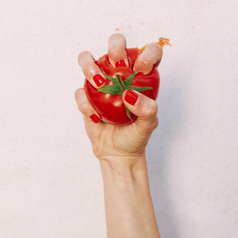 An image of a hand with bright red mani crushing a tomato and an image of a curly haired model covering half of her face with her hand with dark red mani