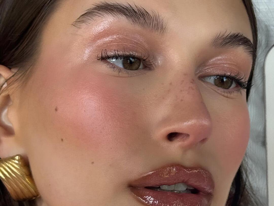 An image of Hailey Bieber sporting a natural makeup look with pink blush, eyeshadow, and lip gloss, complemented by a gold earring