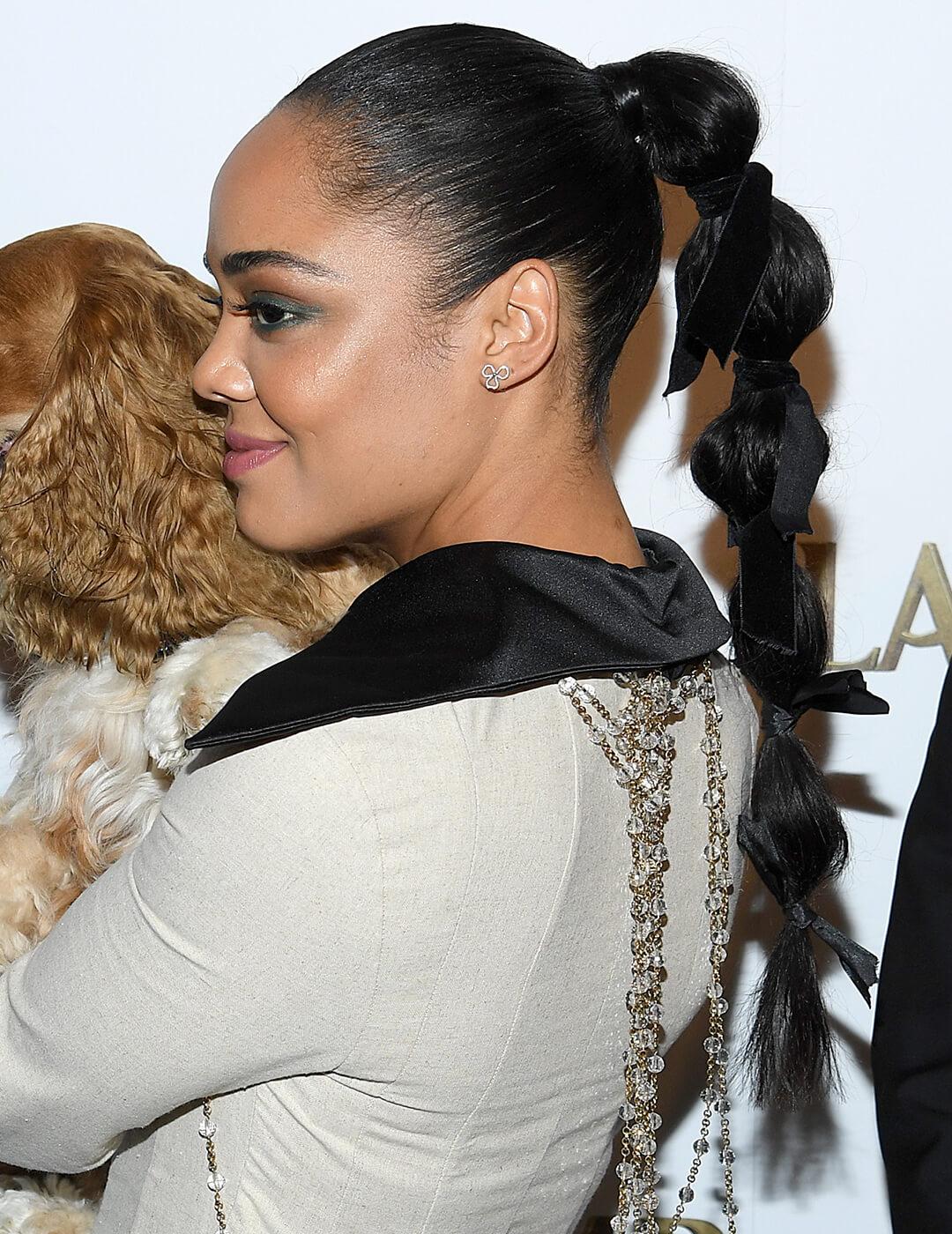 Tessa Thompson wearing a nude outfit with black collar and a bubble ponytail hairstyle with a black bow on each bubble Mobile