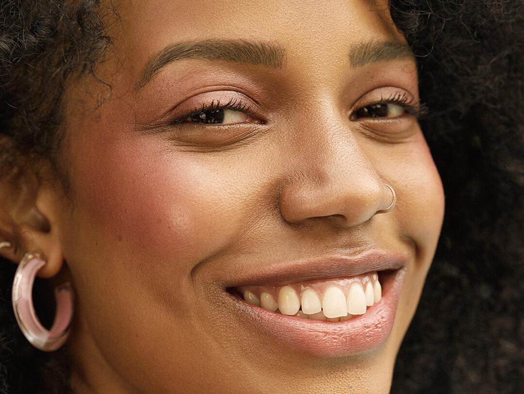 A close-up image of a model wearing rose gold eyeshadow and hoop earrings smiling big