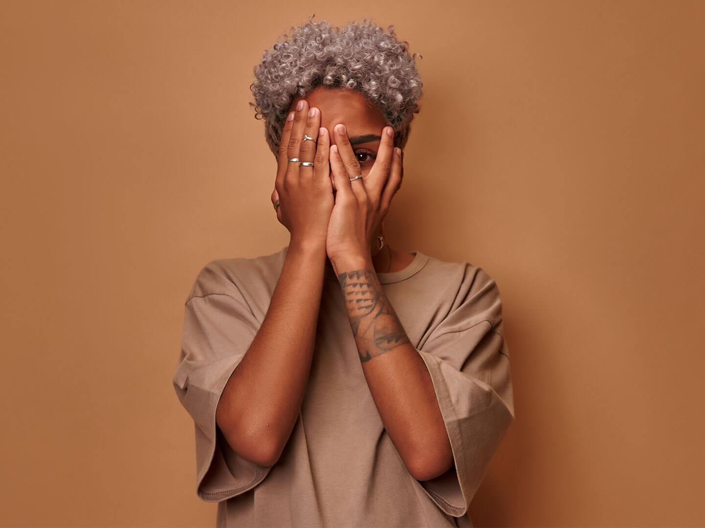 An image of a dark-skinned woman playfully hiding one eye with her hand, glancing out from behind her fingers, against a warm, light brown background