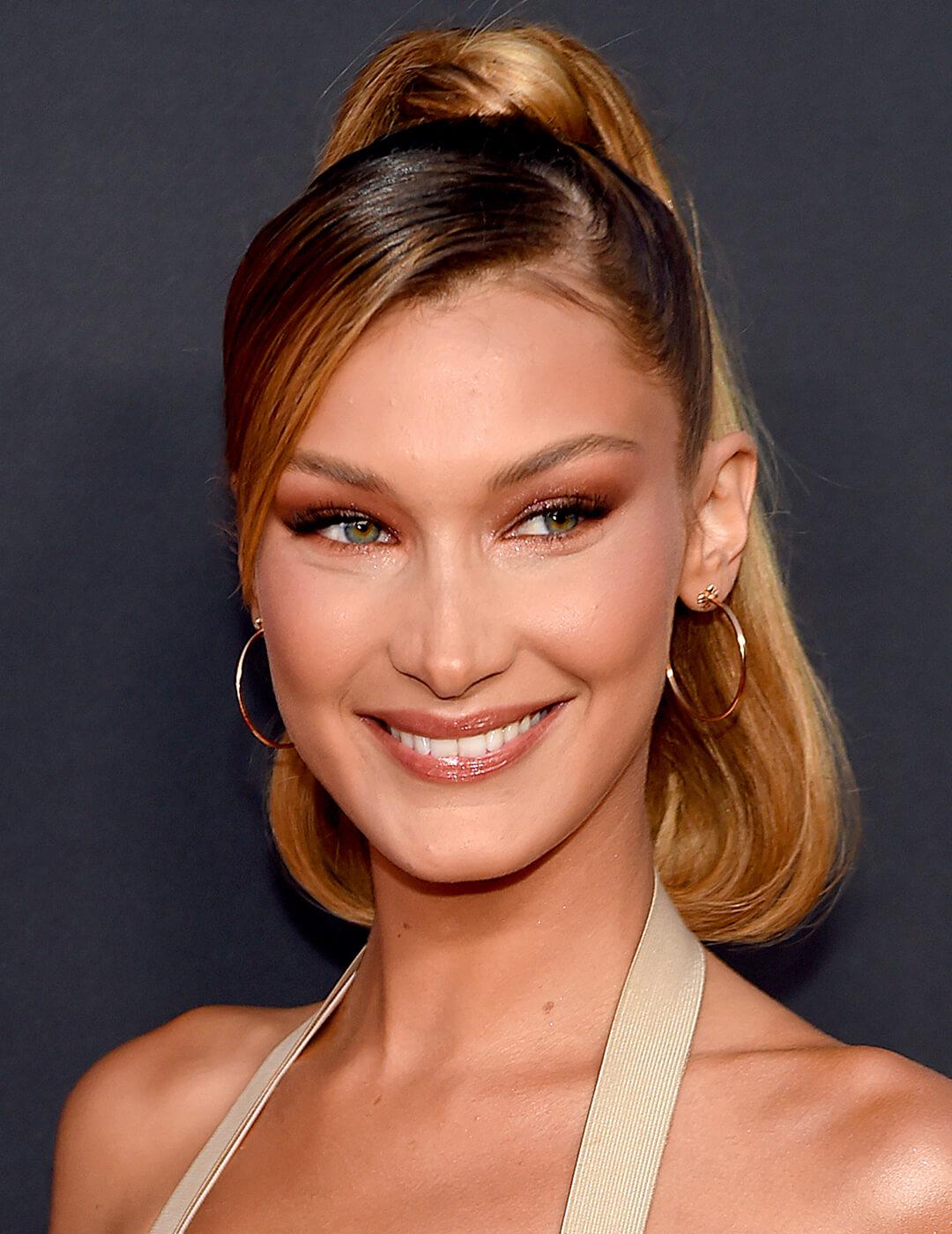 Bella Hadid looking elegant in a gold dress, hoop earrings, rose smoky eye makeup look and sleek, high ponytail hairstyle Mobile