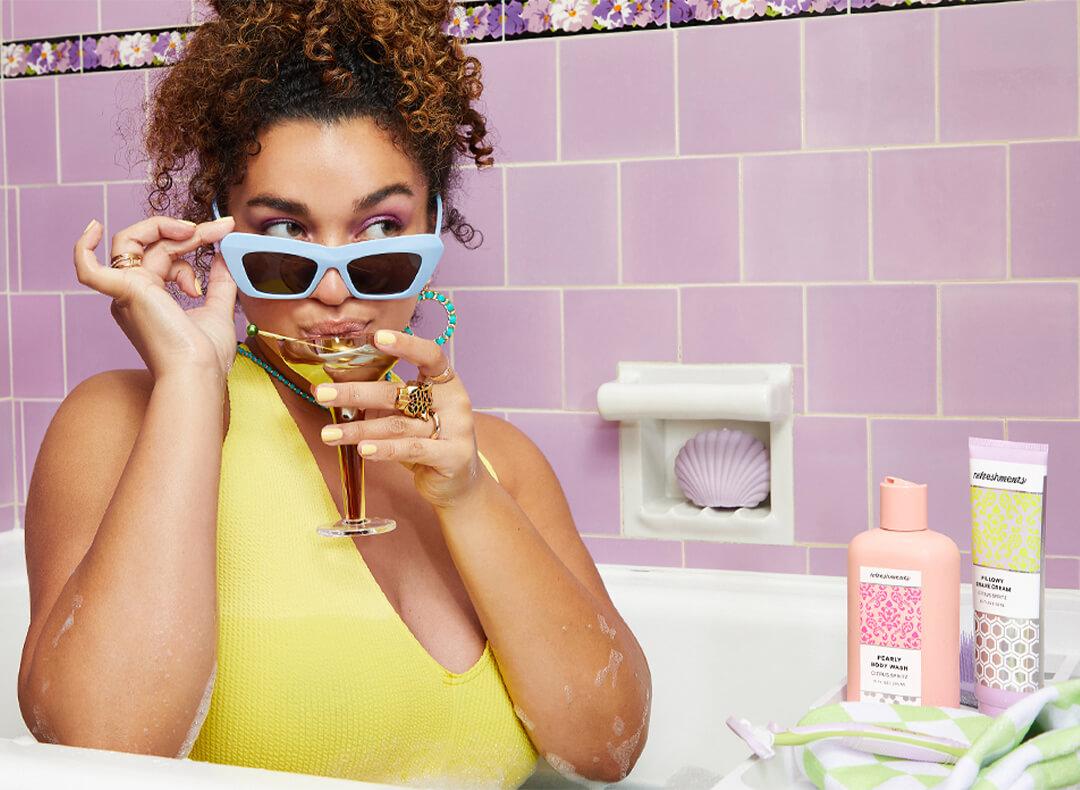 Image of a curly-haired model with sunglasses sipping a cocktail in a bath tub and Refreshments personal care products beside her Mobile