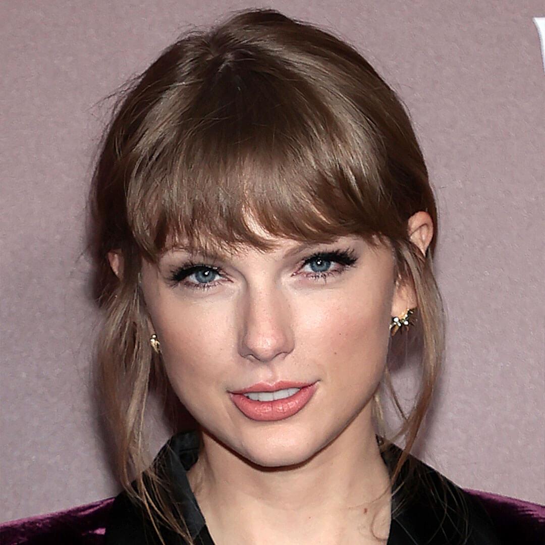 An image of Taylor Swift featuring her Feminine Fluttery Lashes makeup look, gazing directly at the camera while dressed in a velvet suit, her hair is styled with bangs and tied back Mobile