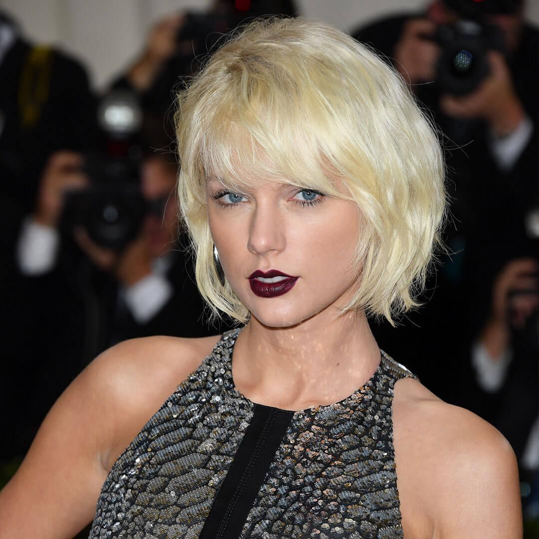 An image of Taylor Swift featuring dark purple-red lips and short blonde hair, dressed in a black sequined tank top dress Desktop