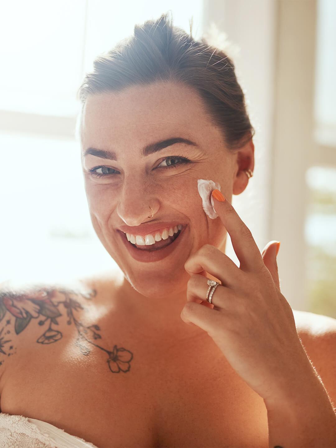 Close-up image of a woman with a shoulder tattoo smiling while applying face cream on her cheek Desktop