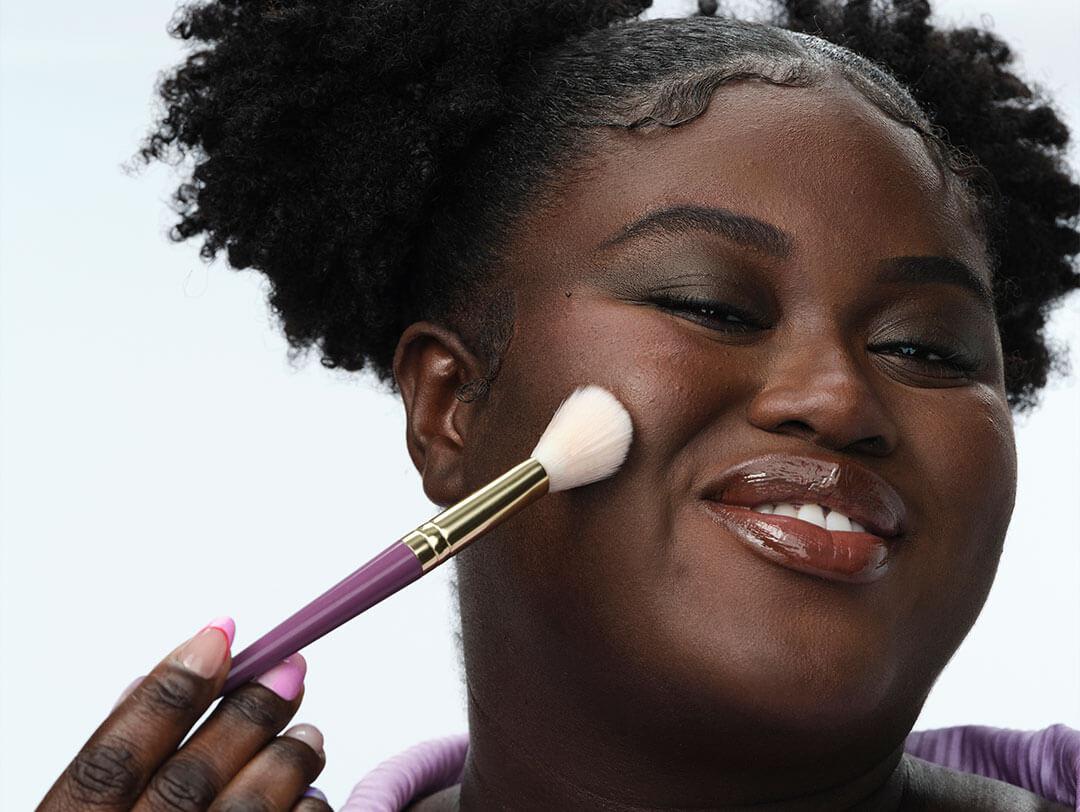 An image of a woman in color, her curly hair styled in a sleek, polished manner, with two buns positioned beside her head while holding a pink makeup brush gently tapping against her cheek Desktop