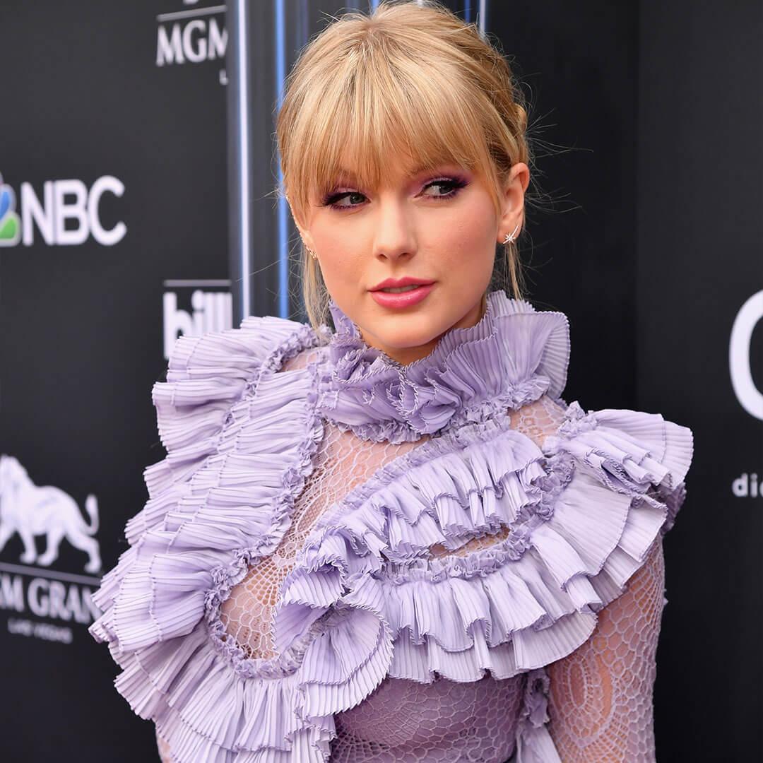 An image of Taylor Swift showcasing her Lavender Haze makeup look, dressed in a ruffled long-sleeve purple dress that features a slightly sheer mesh design Mobile