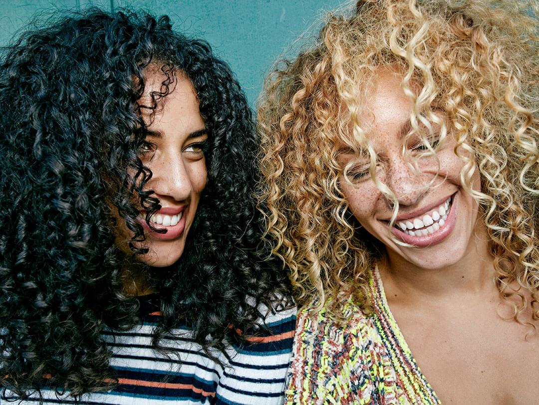 Two women with beautifully textured curly hair, one with dark, tightly coiled curls and the other with blonde, loose curls, showcasing the beauty and vitality of well maintained curly hair Desktop