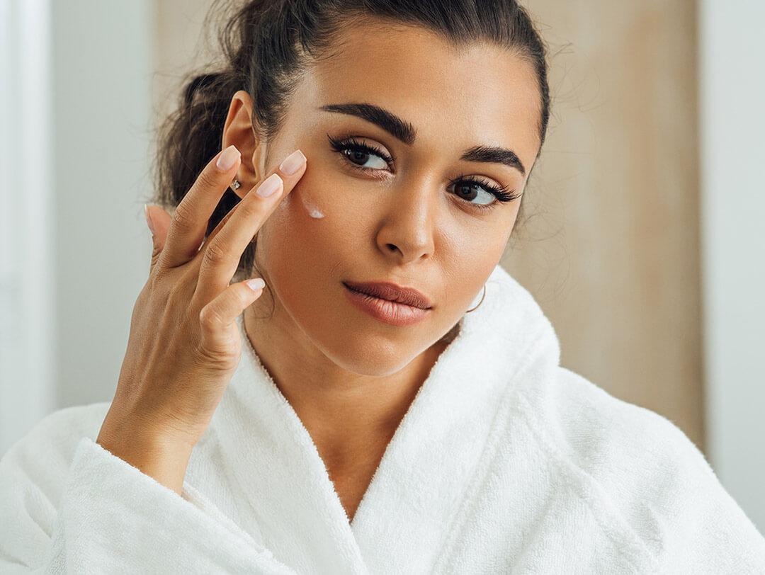 Close-up of a woman in a white robe applying face cream in front of a mirror Desktop