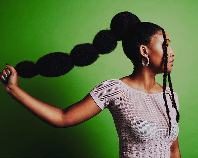 An image of a woman of color with a voluminous braided ponytail against a bright green background, wearing a white textured top and hoop earrings