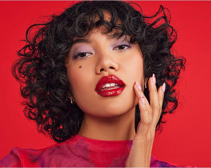 An image of a woman with curly short hair, bold red lips, and lavender eyeshadow poses against a vivid red backdrop, touching her face with polished nails