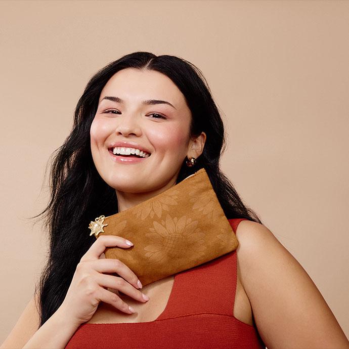 Smiling woman with long dark hair holding a brown floral-textured IPSY Glam Bag, wearing a rust-colored top against a neutral beige background.