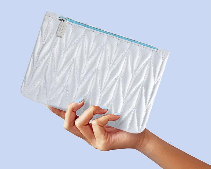 A hand holds a white quilted IPSY Glam Bag against a light blue background. The bag features a geometric stitched pattern, a silver IPSY zipper pull, and a contrasting light blue zipper.