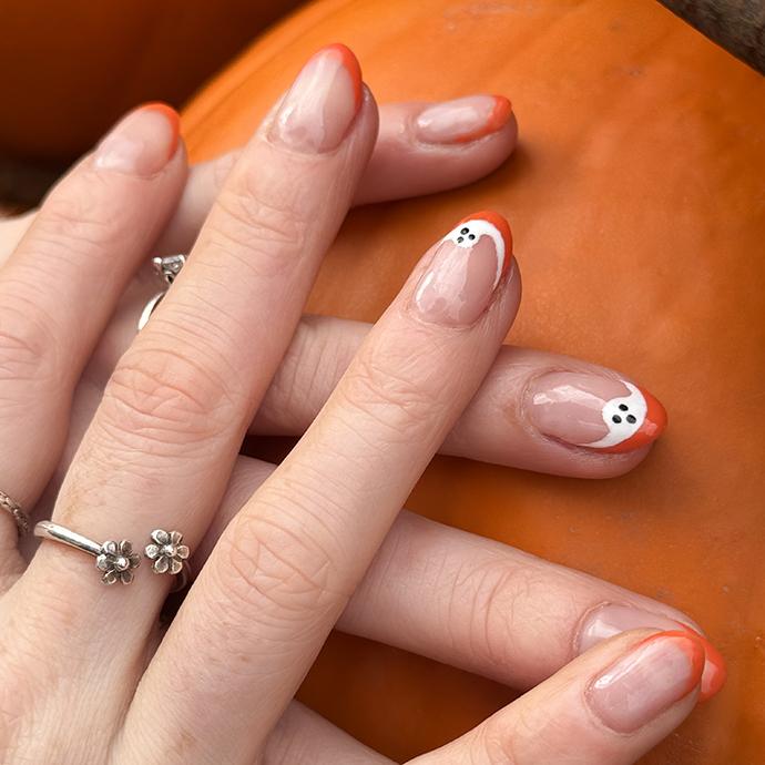 Full frame image of unrecognisable woman holding orange pumpkin displayed on metal hoop, displaying manicured painted nails, Halloween design orange French tip nail art with white ghost, silver ring, focus on foreground