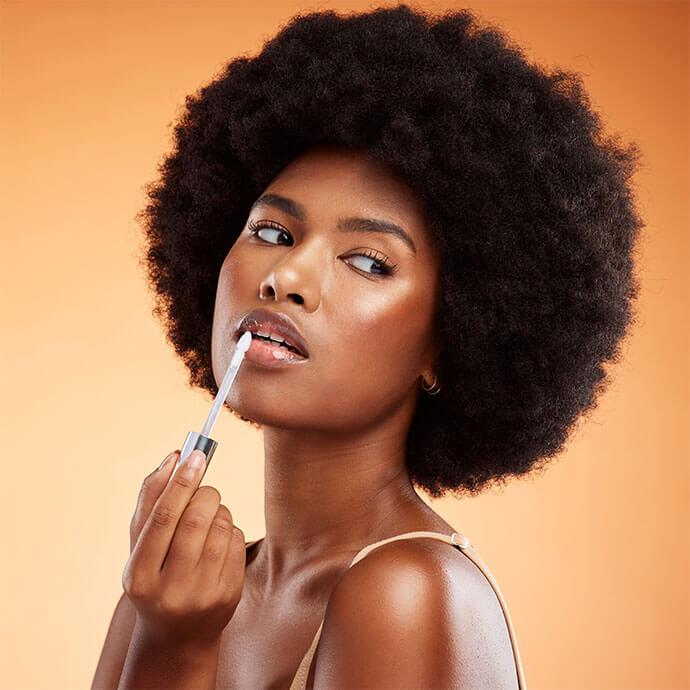Black woman, afro and natural beauty lip gloss treatment for a healthy, shiny and transparent tint, Cosmetics, apply and beautiful face of African model holding makeup tool at orange background