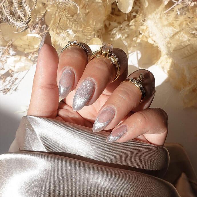 Manicured hand with glossy silver chrome nails is adorned with multiple gold rings against a soft, neutral background