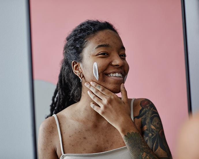 Candid portrait of smiling black woman with acne scars using face cream by mirror