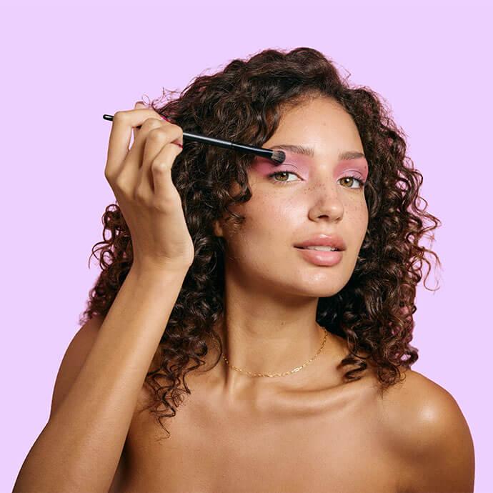 A woman with curly hair applies pink eyeshadow using a makeup brush against a pastel purple background
