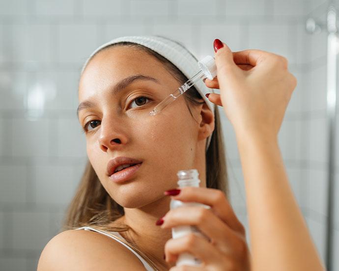 A woman with a white headband and red nail polish applies a clear facial serum to her cheek using a glass dropper in a bathroom