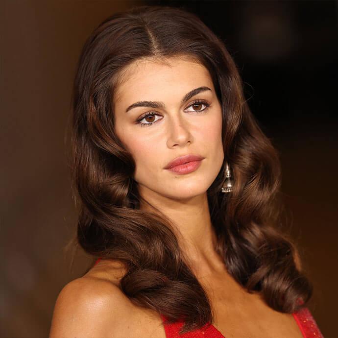 A stylish woman with voluminous, dark brown, center-parted waves and elegant makeup is captured in a headshot