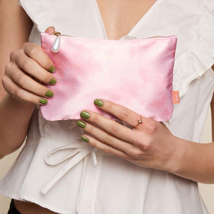 Model holding the April 2026 IPSY Glam Bag, a pink watercolor makeup pouch with a pearl zipper pull.