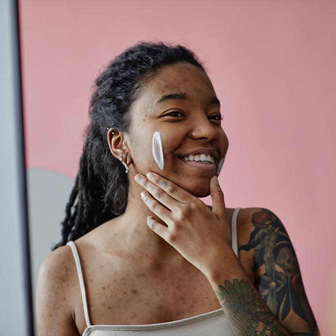 Candid portrait of smiling black woman with acne scars using face cream by mirror