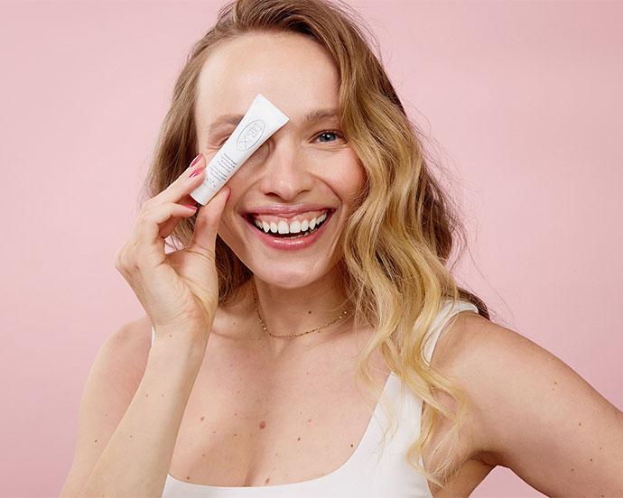 Smiling woman holds a DIEUX SKIN product tube near her eye against a pink background.
