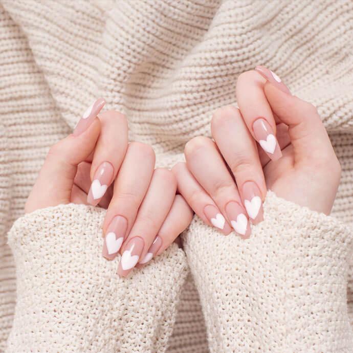 A pair of hands with long nude nails features white heart-shaped tips while resting against a cozy cream knit sweater