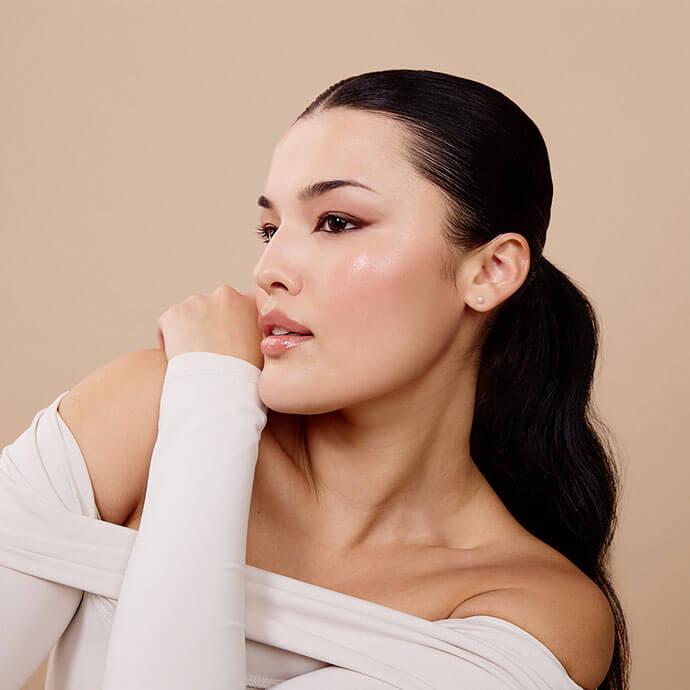 A woman with sleek pulled-back hair poses in profile, showcasing soft, blended eyeliner and a natural glow against a neutral background