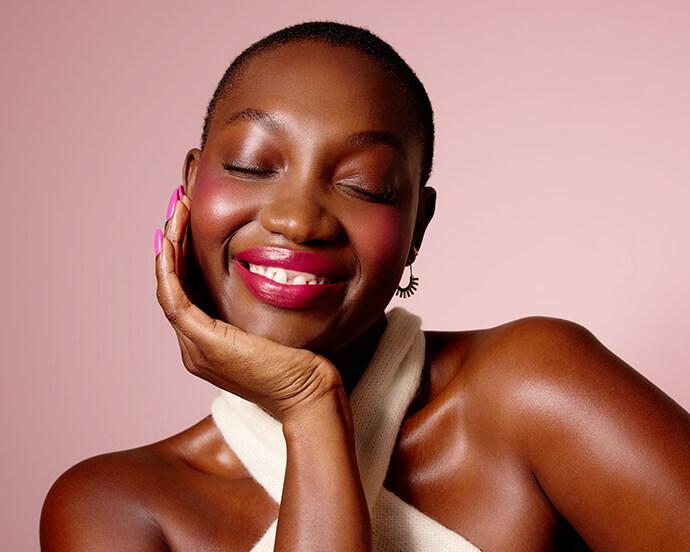 Smiling person with eyes closed and head resting on hand, wearing glossy pink lipstick, soft blush, and shimmery eye makeup against a blush-pink background.