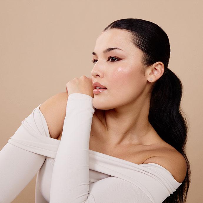Model wearing a sleek low ponytail and soft, glowing spring makeup with rosy blush, glossy lips, and neutral eyeshadow against a beige background.