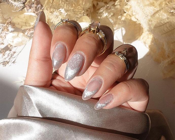 Manicured hand with glossy silver chrome nails is adorned with multiple gold rings against a soft, neutral background