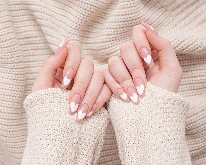 A pair of hands with long nude nails features white heart-shaped tips while resting against a cozy cream knit sweater
