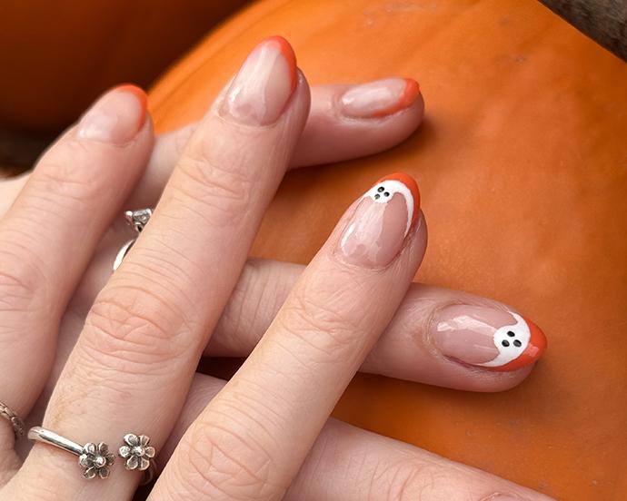 Full frame image of unrecognisable woman holding orange pumpkin displayed on metal hoop, displaying manicured painted nails, Halloween design orange French tip nail art with white ghost, silver ring, focus on foreground