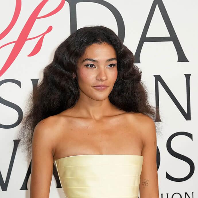An image of Olivia Dean with voluminous, wavy black hair wears a strapless pale yellow dress at a red-carpet event, posing gracefully against a backdrop with bold lettering