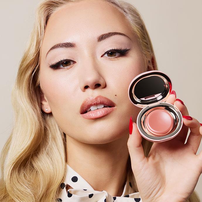 Close-up beauty portrait of a model demonstrating cream blush application, showcasing a soft radiant complexion and holding an open cream blush compact for a step-by-step makeup tutorial.